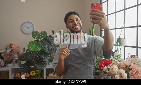 Un homme afro-américain souriant prend un selfie dans une boutique de fleuriste entouré de fleurs, dégageant un esprit convivial et entrepreneurial. Banque D'Images