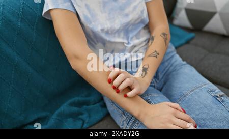 Une jeune femme adulte est assise sur un canapé à l'intérieur, se grattant le bras, présentant des tatouages, une tenue décontractée et du vernis à ongles. Banque D'Images