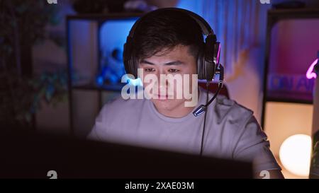 Un jeune homme asiatique portant des écouteurs se concentre intensément dans une salle de jeu sombre et éclairée au néon la nuit. Banque D'Images