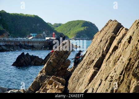 Ville de Gangneung, Corée du Sud - 18 mai 2024 : les pêcheurs grimpent et pêchent depuis la côte rocheuse près du port de Simgok. Le phare rouge et les collines verdoyantes pour Banque D'Images