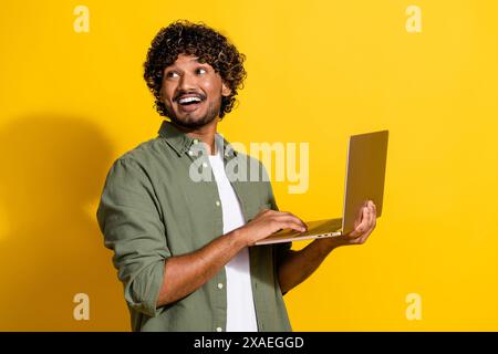 Photo de jeune homme d'affaires aux cheveux frisés en chemise kaki à l'aide d'un ordinateur portable regardant toutes les tâches isolées sur fond de couleur jaune Banque D'Images