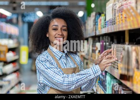 Travailleuse joyeuse portant un tablier, disposant des articles sur une étagère dans une allée de supermarché bien éclairée. Banque D'Images