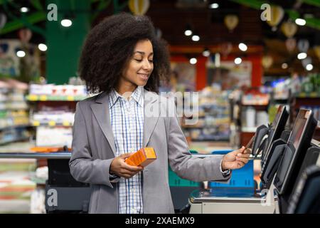 Une femme souriante dans un blazer gris utilise un kiosque libre-service dans une épicerie. Elle tient un produit orange tout en interagissant avec la machine. Banque D'Images