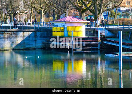 Un petit bâtiment jaune avec un toit rouge se trouve sur un quai à côté d'un plan d'eau. L'eau est calme et le quai est plein de monde Banque D'Images