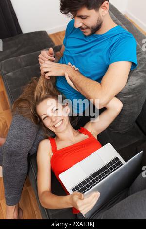 Un jeune couple apprécie le temps de qualité ensemble dans leur salon ; la femme est allongée sur un canapé à l'aide d'un ordinateur portable tandis que l'homme touche amoureusement ses cheveux. Banque D'Images