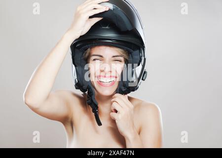 Portrait décontracté et joyeux d'une jeune femme comme elle s'adapte facilement à un casque de moto noir, mettant en valeur la sécurité et la mode avec un sourire vibrant. Banque D'Images