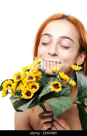 Portrait d'une femme heureuse aux cheveux roux sentant un bouquet de tournesols, rayonnant de joie et de tranquillité sur un fond blanc. Banque D'Images