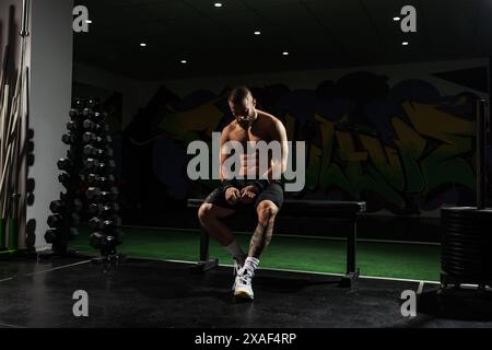 L'homme musclé concentré s'assoit contemplativement dans une salle de sport faiblement éclairée avec des graffitis vibrants. Représente le dévouement et la récupération après l'entraînement. Banque D'Images