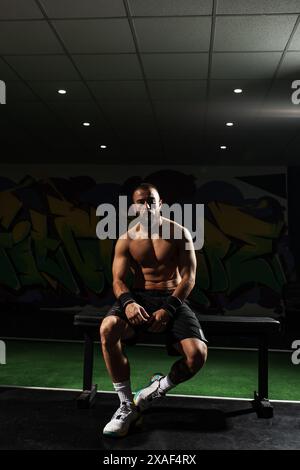 Un athlète masculin concentré et musclé est assis sur un banc, prenant une pause dans une salle de sport moderne avec un fond de graffiti vibrant. Capturer un moment de déterminatio Banque D'Images