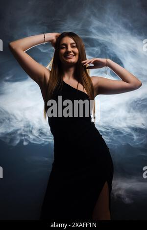 Femme confiante dans une robe noire frappant une pose avec un fond fumé. L'image capture l'élégance, le style et la grâce. Banque D'Images