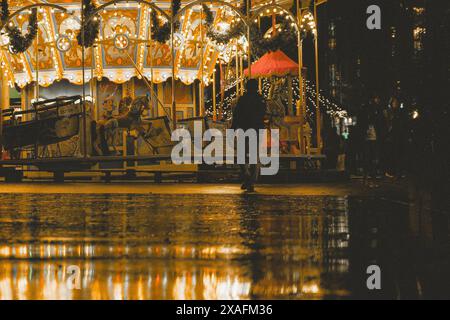 Une personne avec un parapluie rouge marchant à travers un marché de Noël allemand pluvieux, avec un carrousel en arrière-plan. Banque D'Images