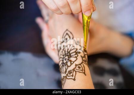 Une image en gros plan montrant une main appliquant soigneusement de la pâte de henné sur un poignet de femme, créant un motif floral magnifique et complexe. L'artiste utilise un Banque D'Images