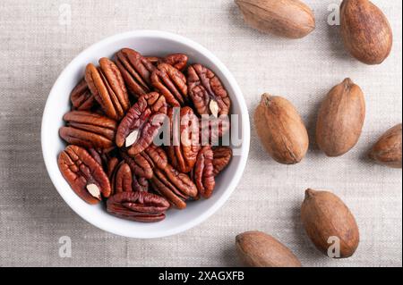 Noix de pécan dans un bol blanc sur tissu de lin. Moitiés de noix de pécan séchées et noix de pécan entières en coquilles, graines et noix de Carya illinoinensis. Banque D'Images