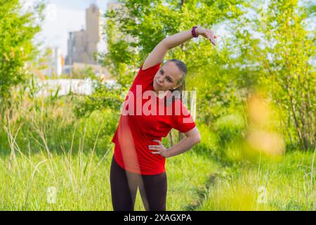 Femme portant une chemise rouge, s'étirant dans un champ herbeux. Elle a son bras droit tendu au-dessus de sa tête et sa main gauche sur sa hanche Banque D'Images