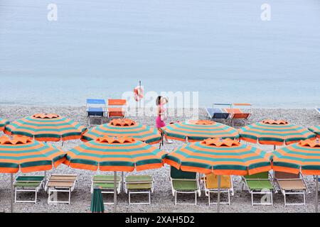 Sur le bord de mer des Cinque Terre à Monterosso al Mare, une femme portant une robe rose passe devant des parasols à rayures vertes et orange sur la plage de galets Banque D'Images