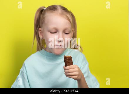 Une belle fille joyeuse aux cheveux blancs de sept ans mange une barre de chocolat sur fond jaune. Les enfants sont des amoureux de la malbouffe sucrée, horizonto Banque D'Images