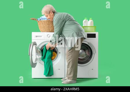 Femme senior avec panier de vêtements mettant le linge dans la machine à laver sur fond vert Banque D'Images