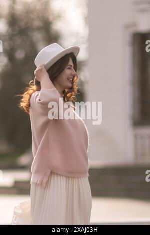 Une femme portant un pull rose et une jupe blanche sourit et tient son chapeau. L'image a une humeur chaleureuse et joyeuse, avec le sourire de la femme et le Banque D'Images