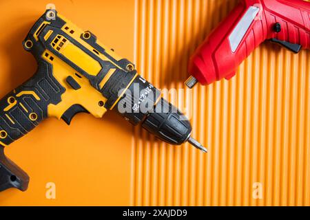 Gros plan d'une perceuse sans fil jaune et noire et d'un tournevis sans fil rouge sur une surface texturée orange. Banque D'Images