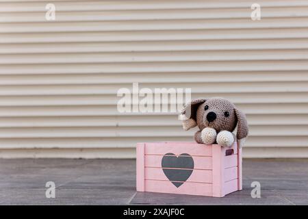 Chiot tricoté dans une boîte rose en bois avec un coeur dessus. Chien jouet crocheté mignon avec sa tête sur ses pattes regardant avec tristesse. Adoptez le concept de ne pas magasiner Banque D'Images