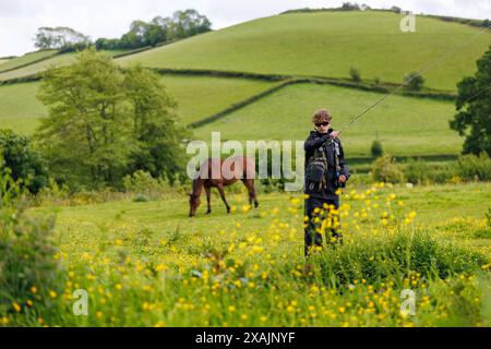 Un adolescent tenant une canne à pêche alors qu'il chasse la truite sur une rive dans le Devon, en Angleterre, en été avec un cheval en arrière-plan. Banque D'Images