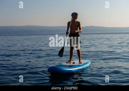 Un jeune adolescent à l'aspect athlétique se tient sur une planche de paddle dans la lumière du soleil doré avec des montagnes en arrière-plan, omis Croatie. Banque D'Images