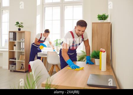 Heureux souriant jeunes nettoyeurs masculins dans le bureau d'essuyage uniforme avec des fournitures de détergent ménager. Banque D'Images