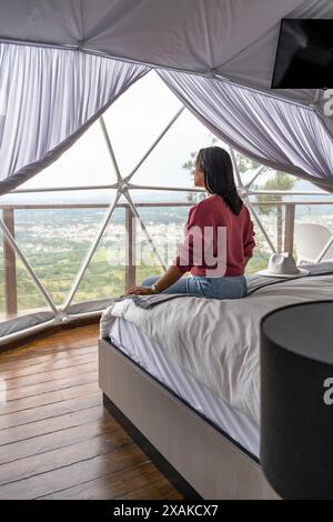 Amérique du Nord, Caraïbes, Île Hispaniola, République dominicaine, Province la Vega, Jarabacoa, Glamping les dômes Jarabacoa, femme assise sur le lit et profitant de la vue Banque D'Images