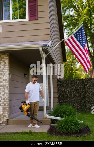 Un jeune homme barbu utilisant un souffleur de feuilles électrique sans fil dans un jardin. Travaux de jardin. Automne, automne jardinage fonctionne dans une cour arrière, sur une pelouse, herbe. Banque D'Images