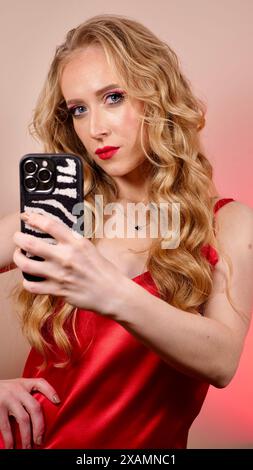 Une jeune femme avec du rouge à lèvres rouge prend un selfie avec de longs cheveux blonds et du maquillage. La fille sourit et regarde la caméra en gros plan Banque D'Images