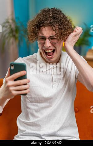 Triste homme caucasien mécontent en utilisant la navigation de clavier de smartphone, perd de devenir surpris soudain résultats de jeu de loterie, mauvaises nouvelles, perte de fortune, échec, délai, virus. Jeune homme aux cheveux bouclés à la maison. Banque D'Images