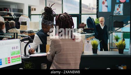 Gars afro-américain ramassant la commande au magasin de vêtements, signant des papiers de confirmation à la caisse enregistreuse. Femme employée préparant des boîtes avec des articles de mode et des accessoires demandés. Caméra B. Banque D'Images