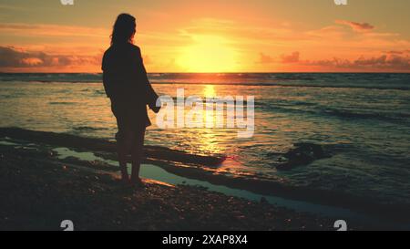 Une personne est debout sur une plage de sable alors que le soleil se couche sur l'horizon, projetant une lumière dorée chaude. Les vagues qui rodent doucement sur le rivage créent une atmosphère paisible. Banque D'Images