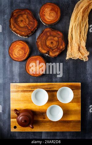 Théière et tasses avec planche à découper en bois et tranches de tronc d'arbre sur fond noir de tableau noir. Pose à plat. Vue de dessus. Banque D'Images