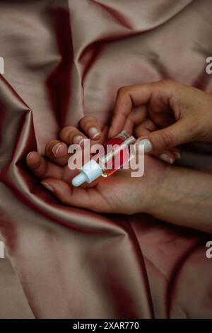 Flacon pipette en verre transparent avec sérum rouge pour le visage, soins du corps dans les mains sur textile rose soyeux. Soins de beauté féminins. Produit esthéticien Banque D'Images