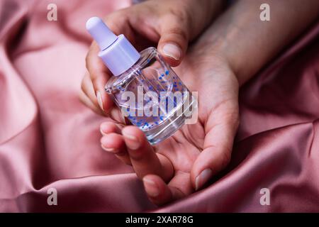 Flacon pipette en verre transparent avec sérum violet pour le visage, soins du corps dans les mains sur textile rose soyeux. Soins de beauté féminins. Produit esthéticien Banque D'Images