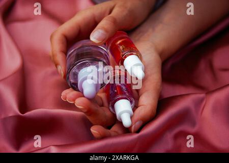 Flacons pipettes en verre transparent avec sérum pour le visage, soins du corps dans les mains féminines sur textile rose soyeux. Soins de beauté pour femmes. Produits esthéticiens. Banque D'Images