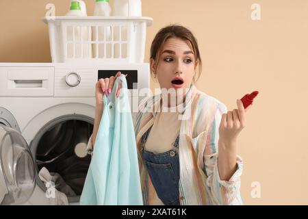 Jeune femme choquée avec du linge sale et du rouge à lèvres près de la machine à laver sur fond beige Banque D'Images