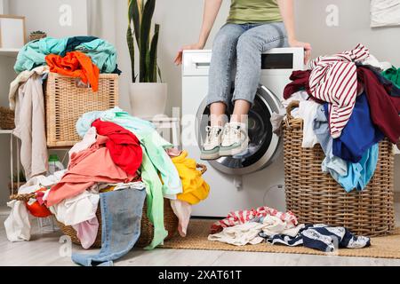 Jeune femme assise sur la machine à laver avec des piles de vêtements sales dans la buanderie Banque D'Images