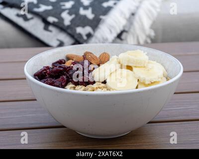 Une vue latérale d'un bol blanc de flocons d'avoine surmonté de tranches de banane, d'amandes et de canneberges séchées sur une table en bois brun, avec une couverture confortable dans le b. Banque D'Images