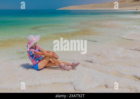 Jeune femme en rose et blanc reposant le long d'une côte de galets avec des eaux vertes claires de la mer morte sous un ciel lumineux et tranquille en Jordanie Banque D'Images