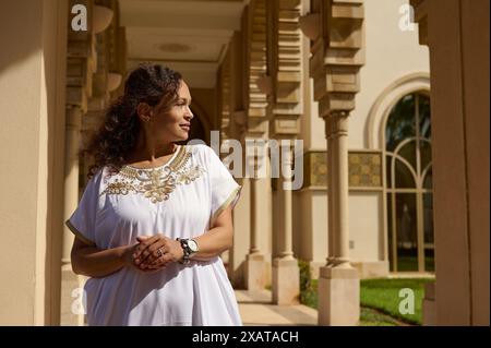 Femme en belle robe traditionnelle blanche se tient gracieusement devant un bâtiment classique soigneusement conçu avec de hautes colonnes. Le moment serein Banque D'Images