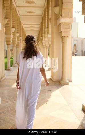 Femme vêtue d'une robe blanche traditionnelle marche à travers un couloir magnifiquement orné, mettant en valeur le patrimoine culturel, l'élégance architecturale. Soleil éclatant Banque D'Images