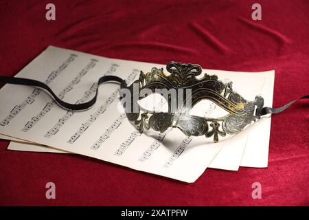 Masque facial élégant et feuilles de musique sur tissu rouge. Représentation théâtrale Banque D'Images