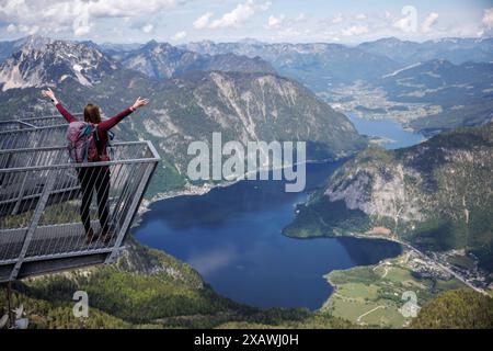 Obertraun, Autriche. 20 mai 2024. Une jeune femme se tient debout avec son sac à dos de randonnée le 20.05.2024, les bras tendus sur l’un des doigts de la plate-forme d’observation à 5 doigts sur le Krippenstein, une montagne de 2108 mètres de haut à la limite nord des montagnes du Dachstein dans la municipalité d’Obertraun en haute-Autriche (Autriche), et donne sur le panorama de montagne. En arrière-plan, le lac Hallstatt avec le village de Hallstatt s’étend au milieu de la vue panoramique sur le paysage montagneux du Salzkammergut haute-Autriche. Crédit : Matthias Balk/dpa/Alamy Live News Banque D'Images