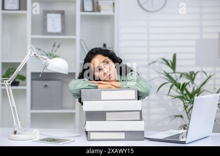 Femme d'affaires fatiguée reposant sa tête sur la pile de dossiers au bureau. Concept surchargé et fatigué dans un espace de travail moderne. Banque D'Images