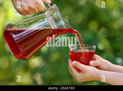 mains d'enfant tenant le verre et le jus de cerise versant de la cruche avec jardin vert sur le fond Banque D'Images