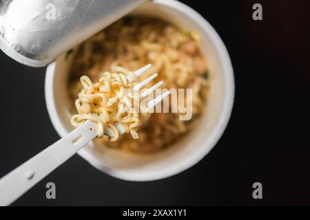 Une tasse de nouilles instantanées avec une fourchette en plastique. Les nouilles sont dans un bouillon avec des morceaux de viande et des légumes. Banque D'Images