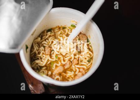 Une tasse de nouilles instantanées avec une fourchette en plastique. Les nouilles sont dans un bouillon avec des morceaux de viande et des légumes. Banque D'Images