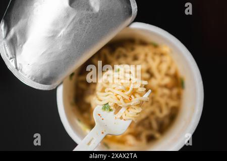 Une tasse de nouilles instantanées avec une fourchette en plastique. Les nouilles sont dans un bouillon avec des morceaux de viande et des légumes. Banque D'Images